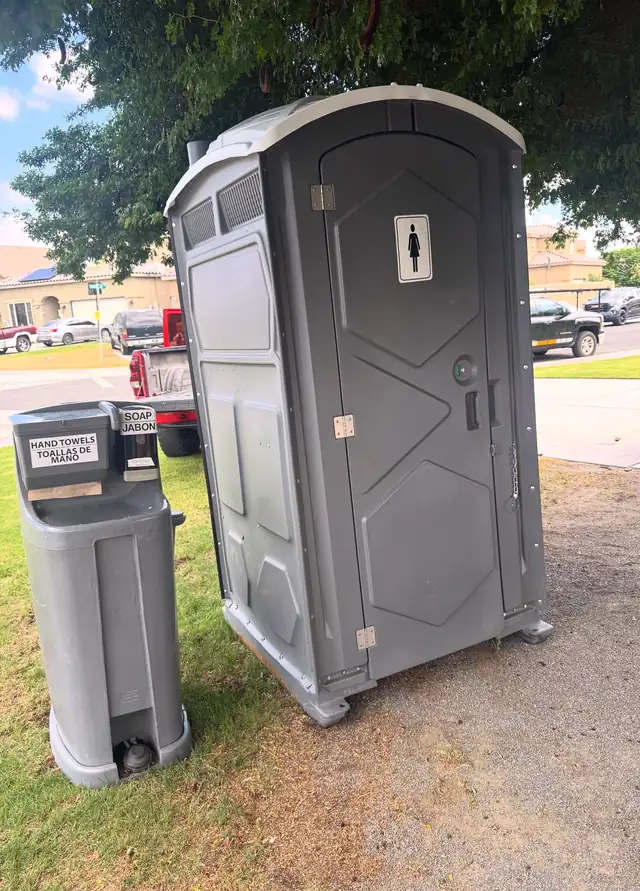 Gray portable toilet next to a freestanding handwashing station on a lawn